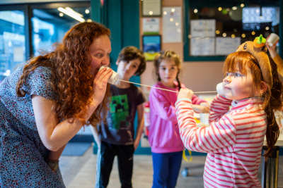 Kirsty speaking into a cup phone and a child listening. Two other children look at the investigation. 