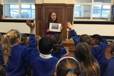 Kirsty presenting to an audience of children with their hands up.