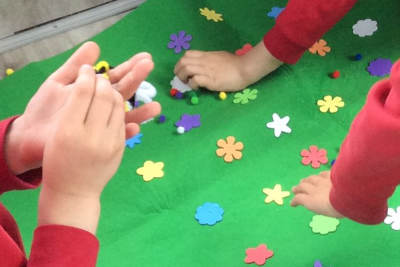 Children's hands holding a pipe cleaner bee, over a green mat with pretend flowers. 