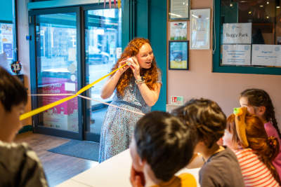 Kirsty is listening into the end of a paper cup, connected by ribbon to other cup phones, where a group of children are speaking and listening. 