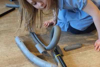 A child is crouched on the floor, looking through their rollercoaster (made from insulating foam pipe, elastic bands, and plastic tubes) to see if their marble got through. 