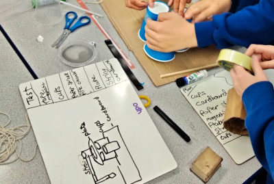 Children building using cellotape, cups and cardboard, whilst their whiteboard labelled diagram can be seen in the foreground.